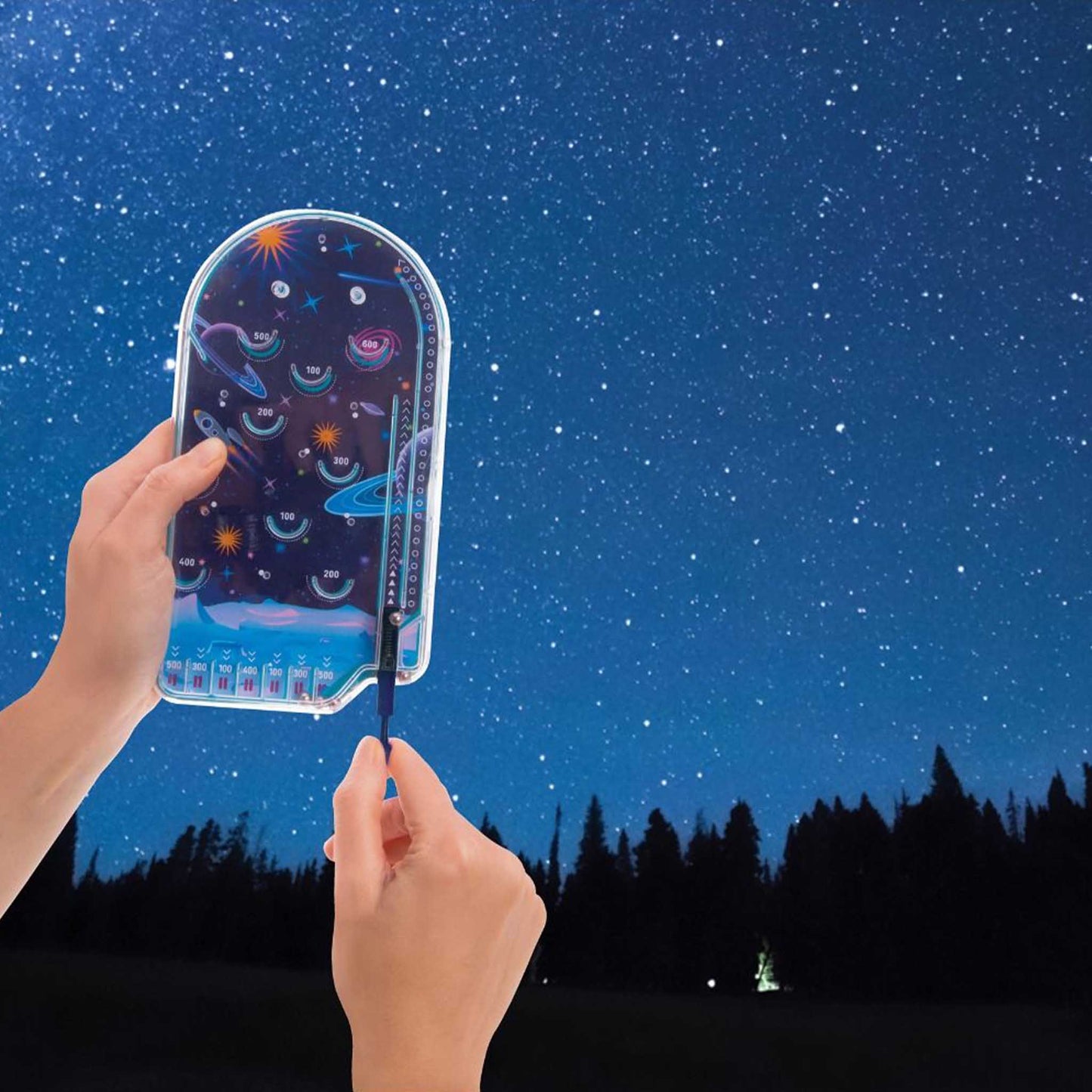 Hand holding a star themed pinball game against a starry night sky with trees in the background