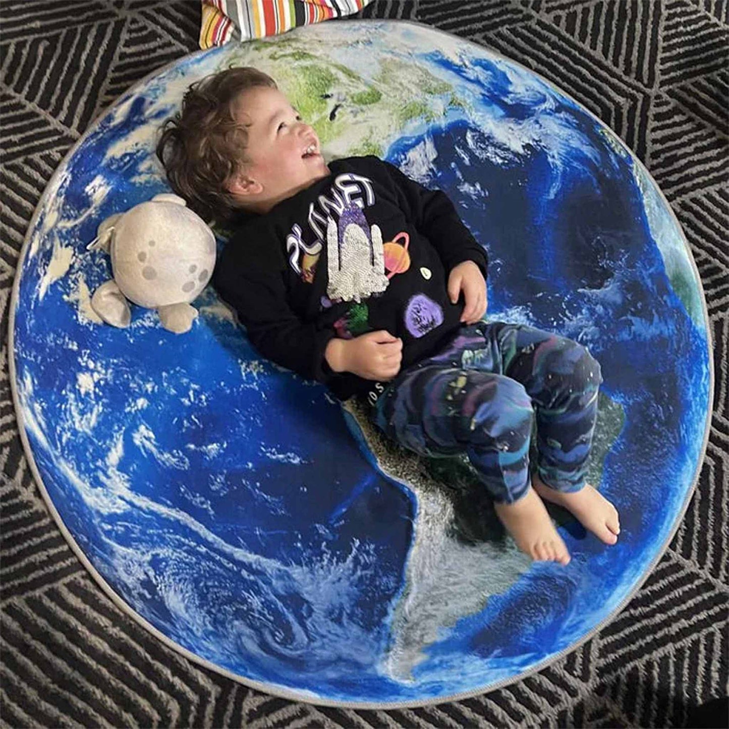 Child lying on a large地球-shaped cushion with a toy, on a patterned rug.