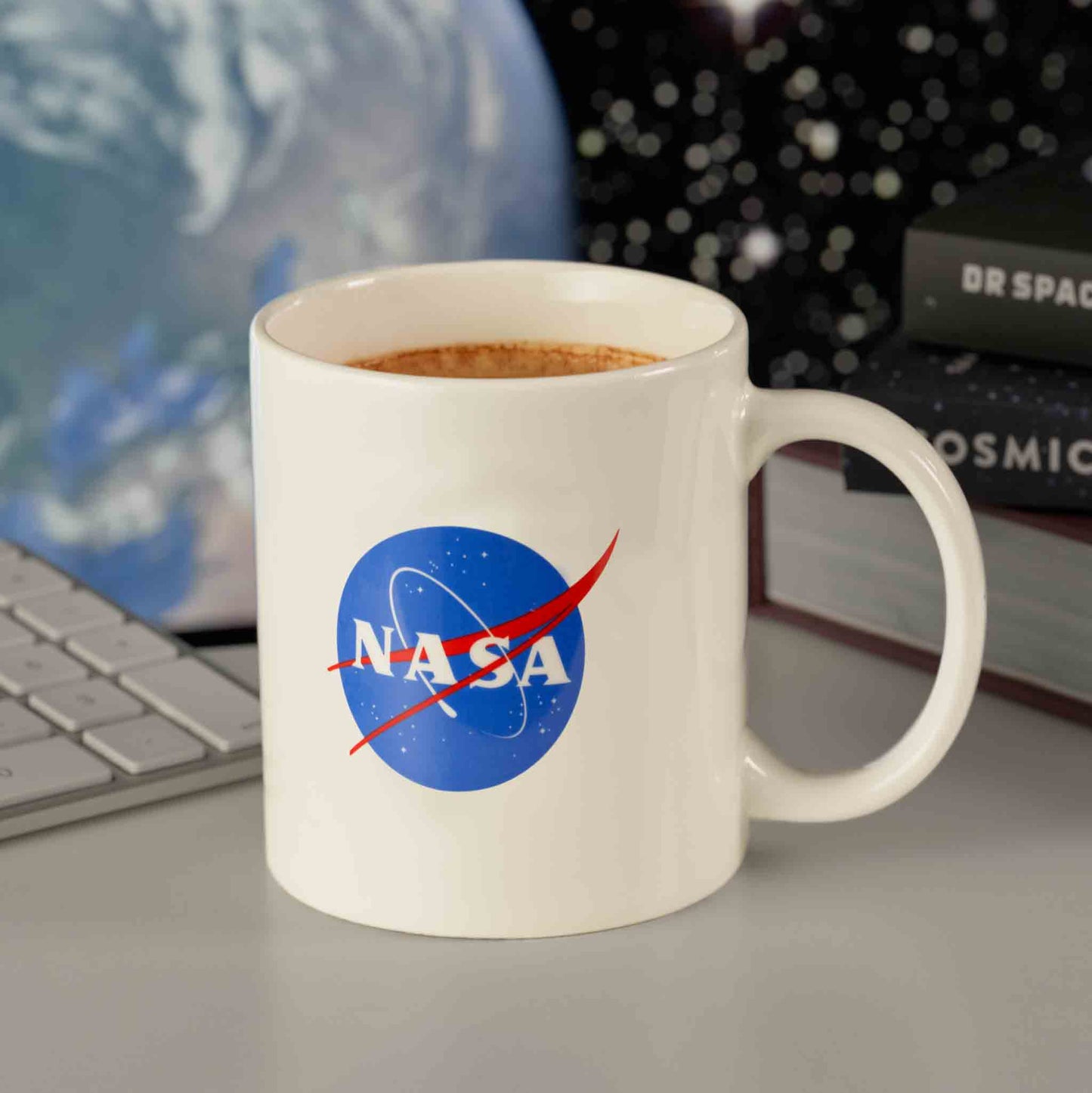 White mug with NASA logo on a desk with books and a laptop in the background