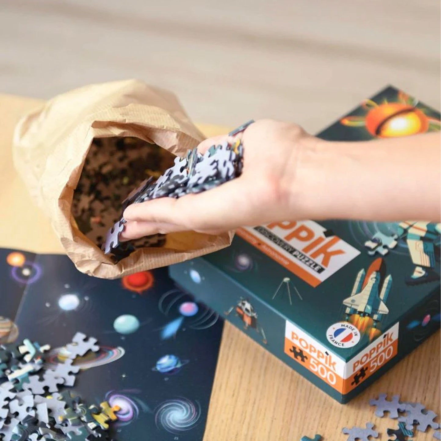 Hand reaching into a paper bag filled with puzzle pieces next to a Popik Puzzles box.